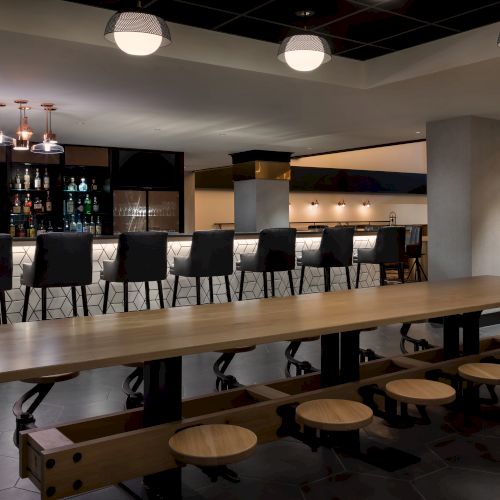 A modern bar interior with pendant lights, a stocked liquor shelf, stools, and glassware behind the counter, with a lounge seating area.