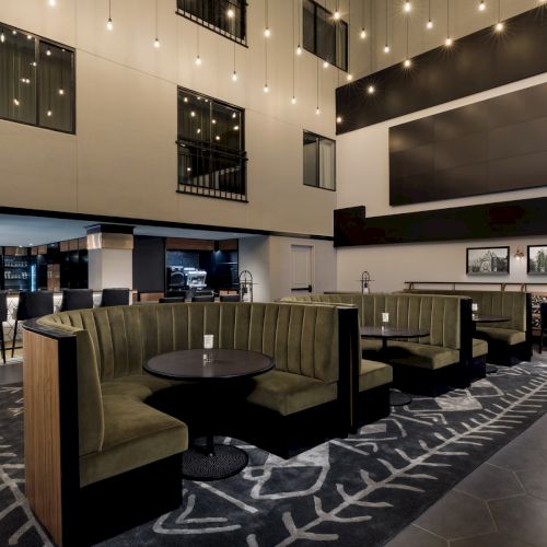A modern hotel lounge with green booth seating, round tables, pendant lights, a hallway bar, and framed artwork on the walls.