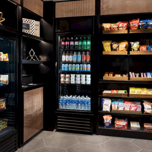 A hotel-style pantry with refrigerated drinks, snacks, and boxed food in illuminated shelves and glass-door fridges.