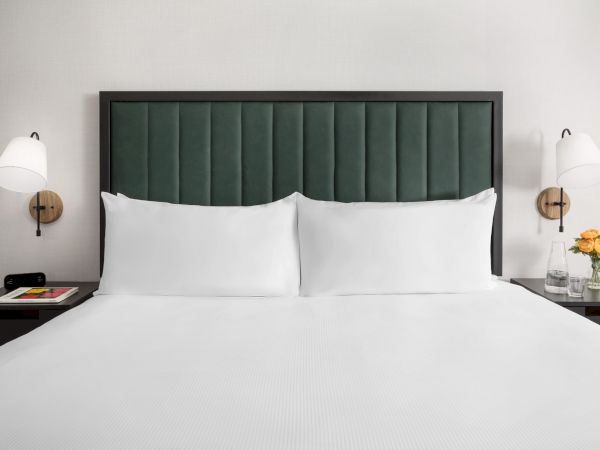A neatly made hotel bed with a dark green padded headboard, two white pillows, white bedding, and matching nightstands with lamps on each side.