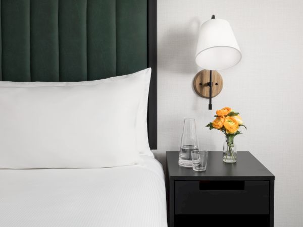 A neatly made bed with white linens and a dark green headboard, beside a black nightstand holding a lamp, flowers, and glassware.