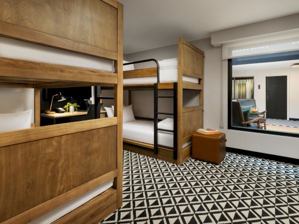 A hotel room with wooden bunk beds, a geometric black-and-white floor, and a separate seating area visible through a large window.