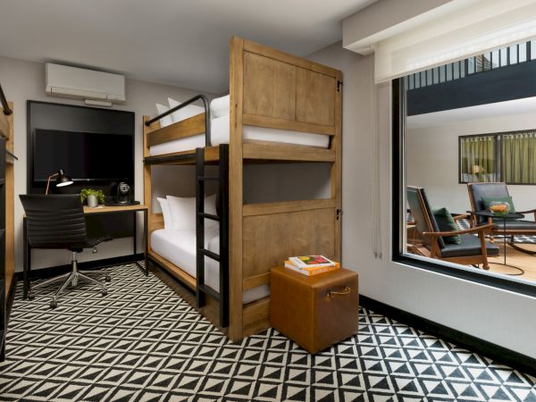 A modern room with a wooden bunk bed, a small desk and chair, a suitcase stool, and a patterned black-and-white floor, with a mirrored wall.