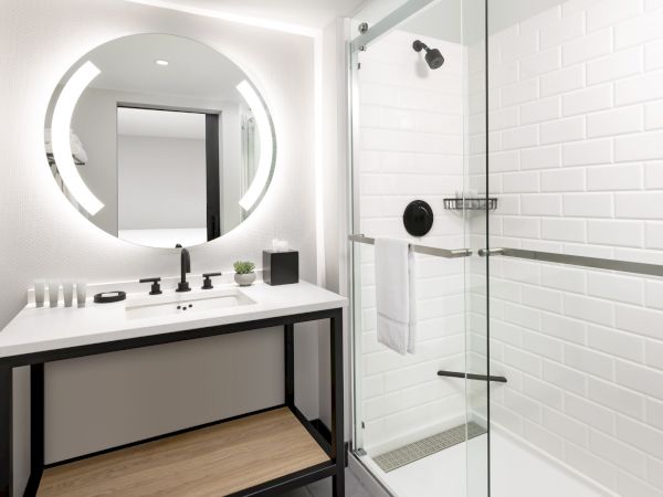 Modern bathroom with a round backlit mirror, white vanity, black hardware, and a glass-walled shower with white subway tiles. It&rsquo;s clean and contemporary.