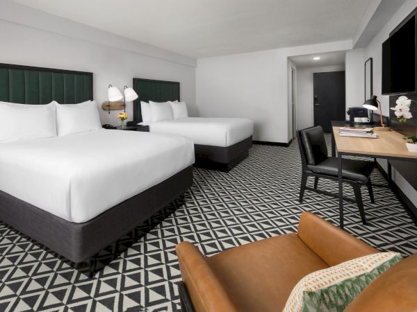 Two queen beds with white linens, a patterned floor, a seating area, and a desk in a modern hotel room. The decor is black, white, and green accents.