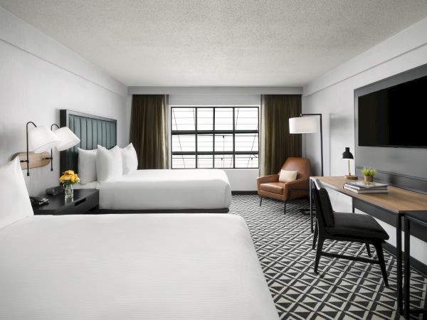 A modern hotel room with two white beds, a seating area by the window, a desk and chair, wall-mounted TV, and a geometric patterned floor, bright and clean.