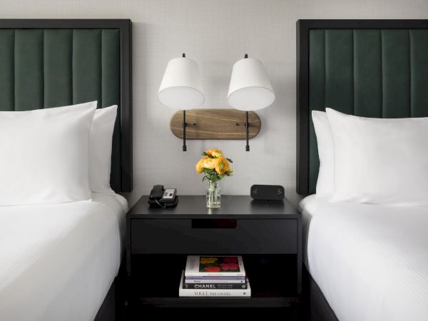 Two neatly made hotel beds with a shared nightstand between them, a small flower vase, books, and wall-mounted lamps.