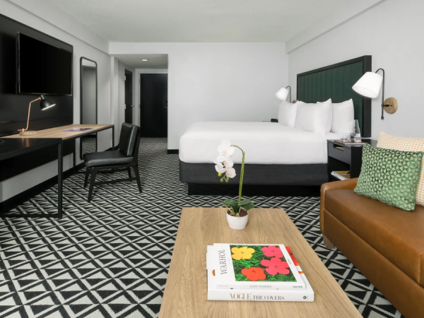A modern hotel room with a king bed, desk, patterned rug, coffee table, and a brown leather sofa with pillows.