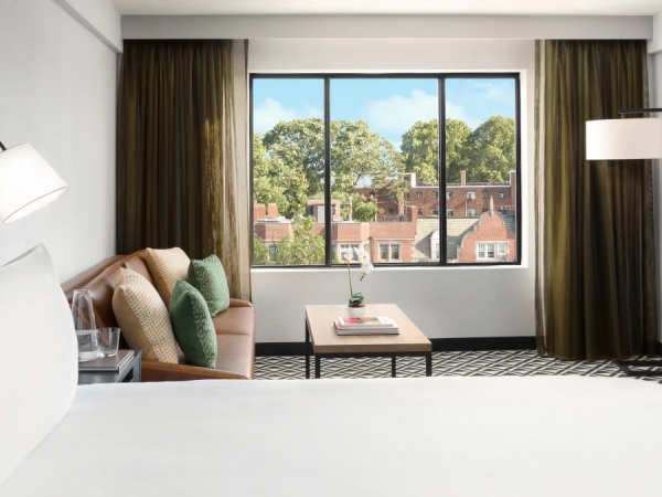 A modern hotel room with a plush bed, seating area, large window overlooking a town, floor lamps, and a cozy, bright vibe.