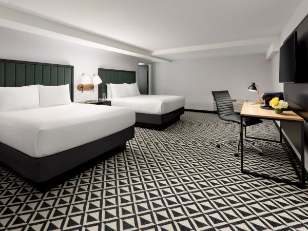 A modern hotel room with two white beds, a work desk with chair, patterned carpet, and two wall-mounted TVs.