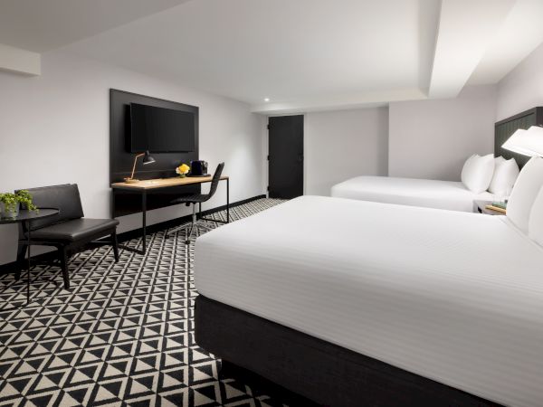 A modern hotel room with a large bed, TV at a desk, an office chair, and patterned carpet with a bedside lamp.