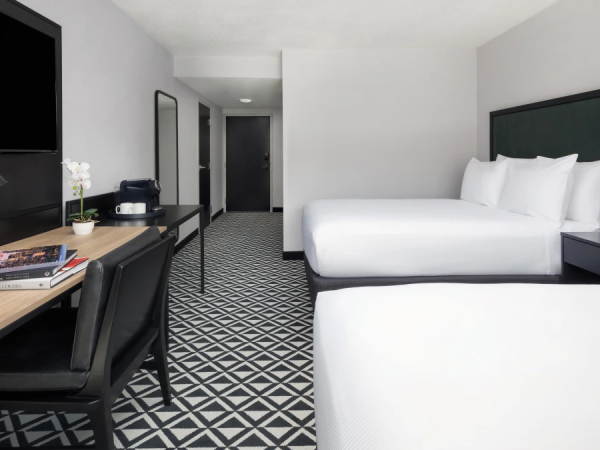 A hotel room with two white beds, a desk and chair, a wall-mounted lamp, a flat-screen TV, a mirror, and a geometr patterned floor; a hallway ahead.