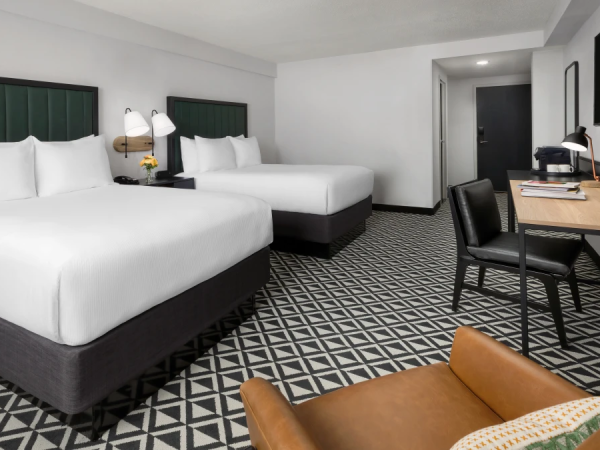 Two double beds with green upholstered headboards, a work desk and chair, patterned carpet, and a seating area in a modern hotel room.