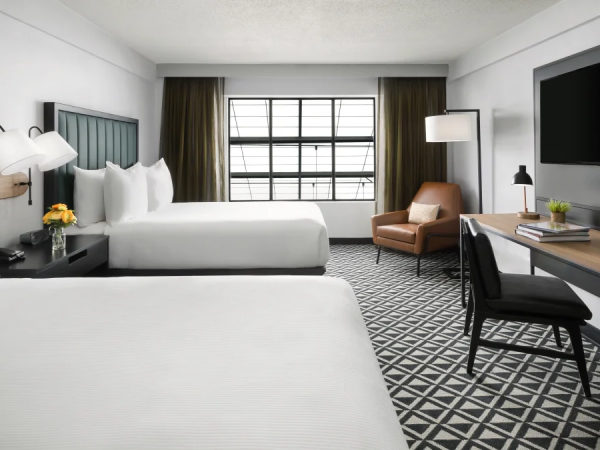 Two queen beds in a bright hotel room with a desk, chair, flat-screen TV, and a window seating area; neutral tones and geometric carpet.