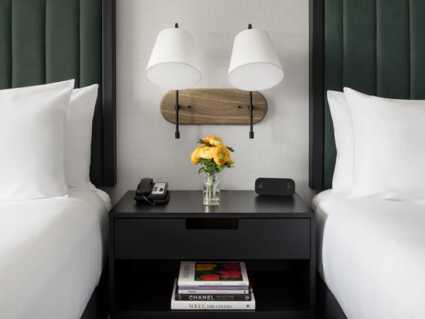 Two neat twin beds with white linens flank a dark nightstand, featuring a vase of yellow flowers, wall lamps, and a stack of magazines beneath.