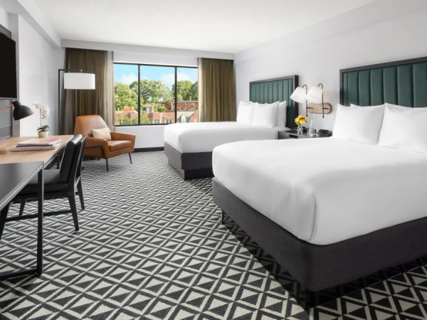 A modern hotel room with two plush queen beds, a work desk, a flat-screen TV, a seating area, and a large window showing trees outside.