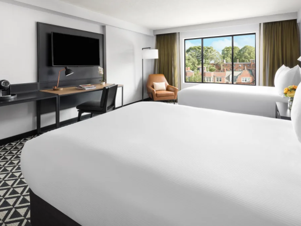 A modern hotel room with two large white beds, a desk setup with a monitor, a chair, a flat-screen TV, a window view, and patterned carpet.