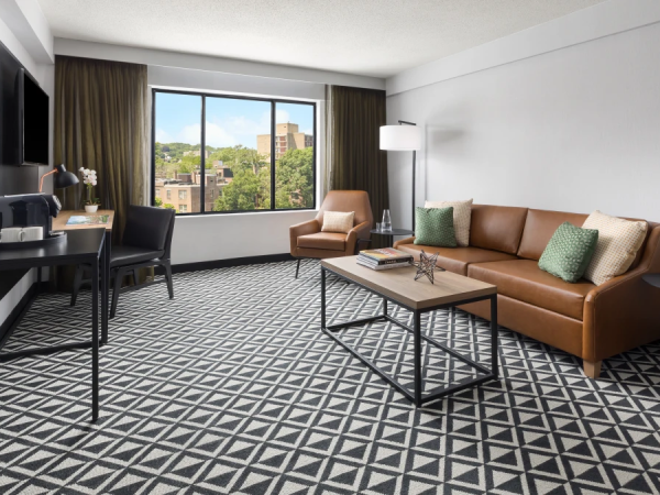A modern hotel living room with a brown leather sofa, geometric carpet, coffee table, large window view, and a desk area with chairs.