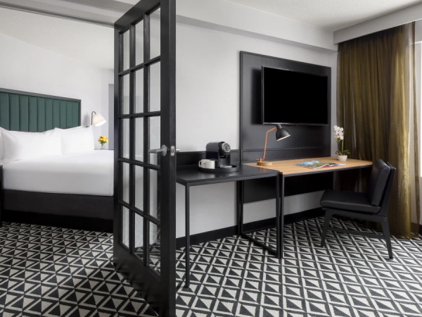 A modern hotel room with a separate sleeping area, glass divider, desk and chair, wall-mounted TV, patterned floor, and large window with a city view.