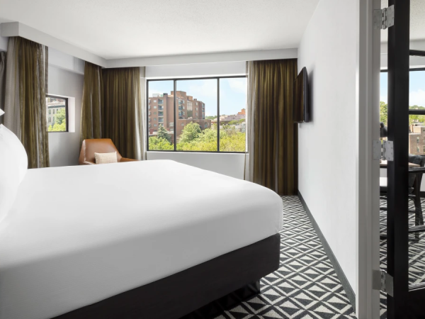 A modern hotel room with a large bed, white linens, floor-to-ceiling windows, a patterned floor, a balcony door, and a view of trees and buildings outside.