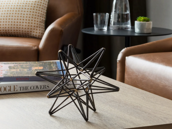 A cozy living room with brown leather chairs, a wooden coffee table, stacked magazines (Vogue and others), and a geometric metal sculpture.