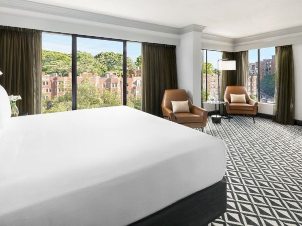 A modern hotel room with a large bed, geometric carpet, two armchairs by the window, and big glass doors opening to a city view.