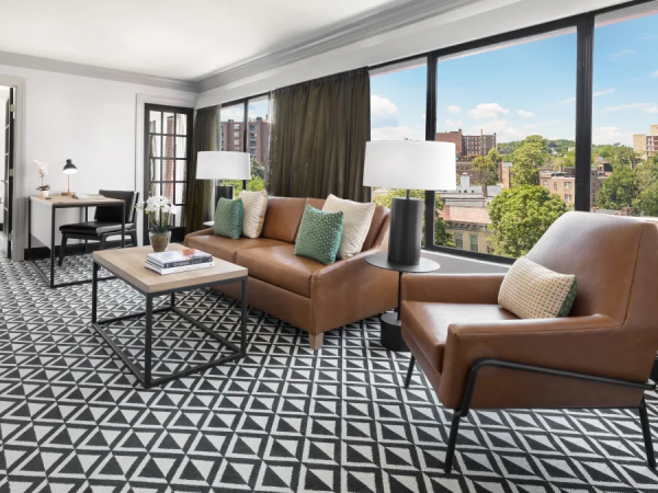 A modern living room with large windows, brown leather sofa and chairs, geometric black-and-white carpet, coffee table, lamps, and city views, stylish and bright.