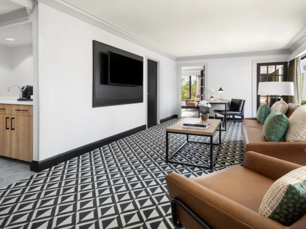 A modern hotel suite with a patterned carpet, a wall-mounted TV, a kitchenette area, and a living room with brown leather chairs, a coffee table, and a sofa, bright with natural light.
