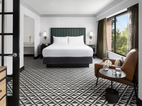A stylish hotel room with a king bed, geometric patterned carpet, a small seating area, large window view, and modern furnishings&mdash;ending in a sentence.