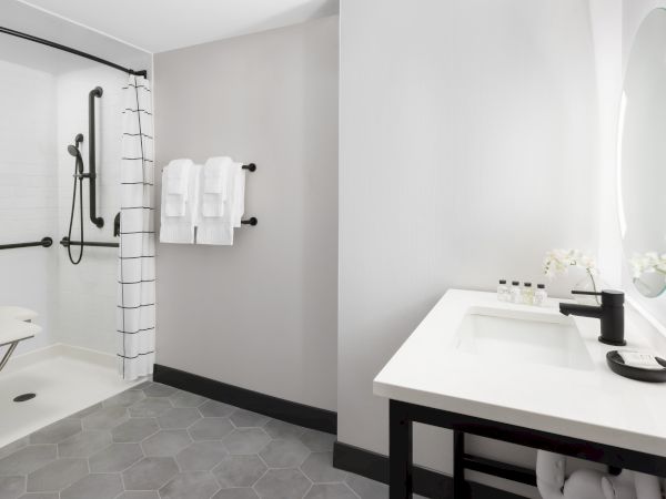 A bright modern bathroom with a walk-in shower, striped curtain, folded towels, a white sink, and toiletries by a large mirror.