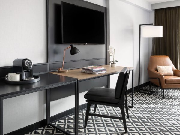 A modern hotel room featuring a work desk with a coffee station, a wall-mounted TV, a chair, and a reading lamp by a large window.