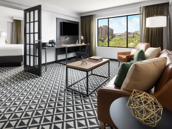 A modern hotel suite with a living area and workspace, patterned floor tiles, a brown leather sofa, coffee table, large window view, and a separate bed area.