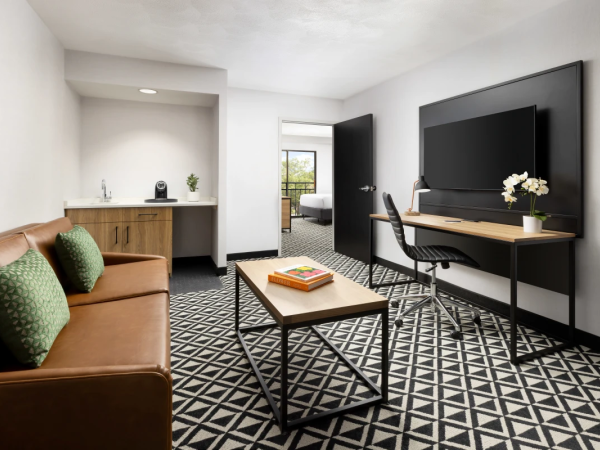 A modern hotel suite with a patterned black-and-white floor, brown leather sofa, wood desk, wall-mounted TV, and a small kitchenette, plus a coffee table and doorway to a bedroom.