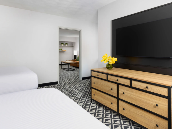 A bright hotel bedroom with two white beds, a wooden dresser under a mounted TV, yellow flowers, and a doorway leading to another room with a patterned carpet, all in a modern, minimal style.