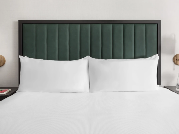 A neatly made hotel bed with a dark green padded headboard, two white pillows, white sheets, and matching bedside tables with lamps and a small vase of flowers on the right.