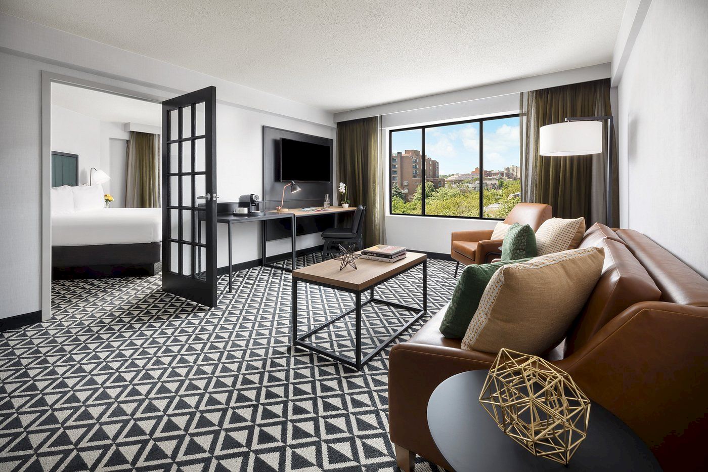 A modern hotel suite with a patterned floor, living area, TV, desk, and a connected bedroom visible through a glass door.