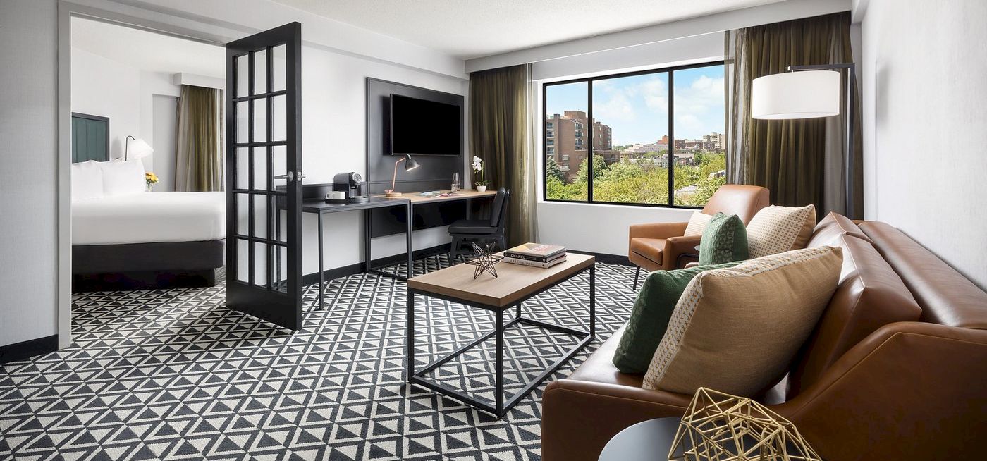 A modern hotel suite with a patterned floor, living area, TV, desk, and a connected bedroom visible through a glass door.