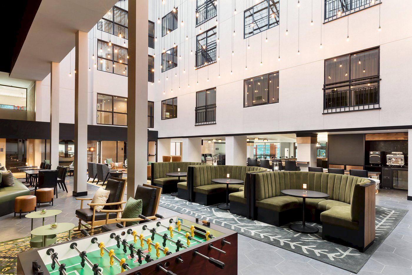 A modern hotel lobby with white walls, comfy green sofas, and a foosball table in a bright, open seating area.