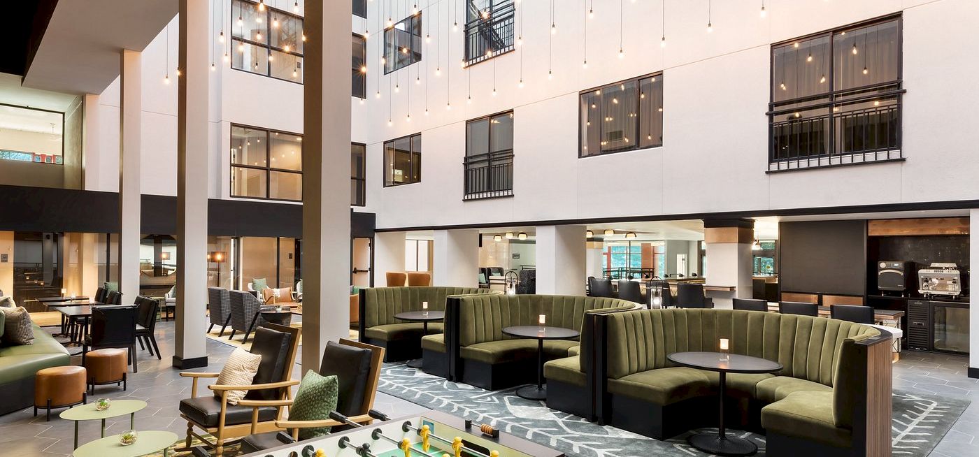 A modern hotel lobby with white walls, comfy green sofas, and a foosball table in a bright, open seating area.