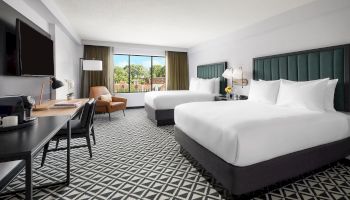A modern hotel room with two large white beds, a desk, seating area, patterned carpet, and a sunny window view.