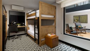 A modern dorm room with a wooden bunk bed, a small desk area, a black-and-white geometric floor, and a large window looking into a lounge area.