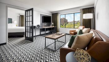 A modern hotel suite: living area with a sofa, coffee table, and patterned floor; a desk, TV, and large window view; doorway to a bedroom.