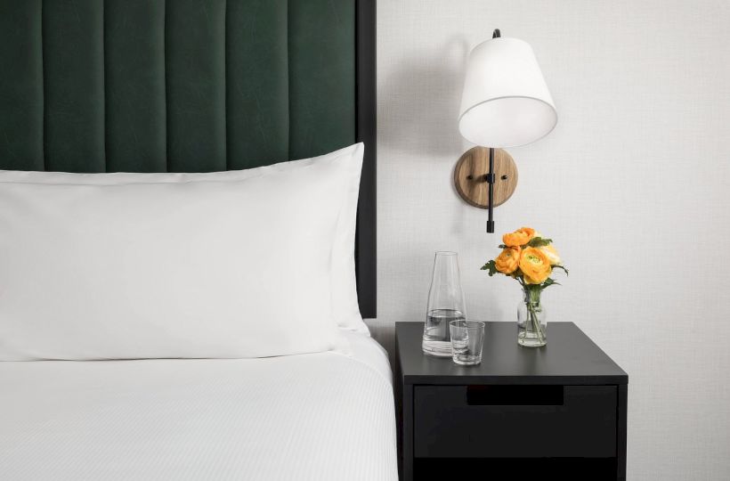 A modern bedroom scene with a neatly made bed, a dark nightstand, a white wall lamp, and a small vase of yellow roses beside a glass of water.