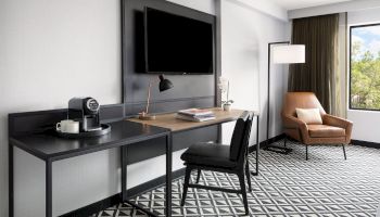 A modern hotel room with a desk, chair, wall-mounted TV, coffee machine, lounge chair, floor lamp, and a geometric patterned carpet, near a window.