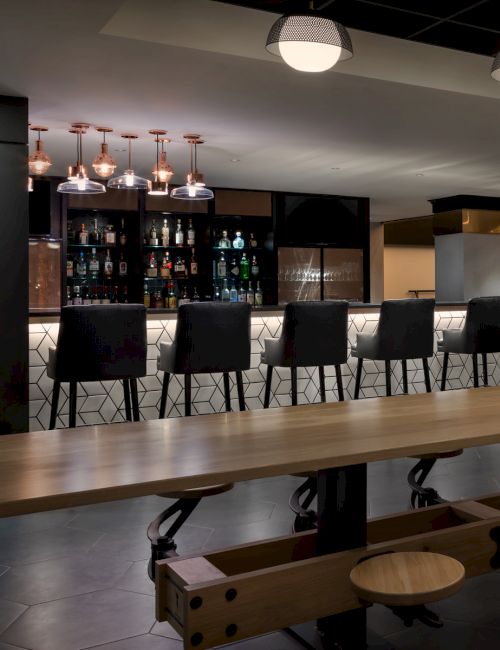 A modern bar or restaurant interior with a long wooden communal table, bar stools along the back counter, and warm pendant lighting above.