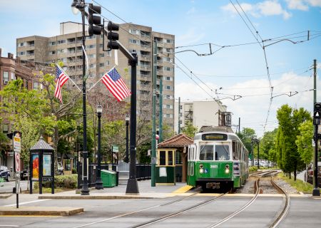 A vintage green tram on tracks in a city square, with flags, trees, a bus stop, and tall buildings in the background, under a blue sky.