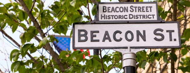 A street sign reading "BEACON ST." with a smaller banner above saying "BEACON STREET HISTORIC DISTRICT," set among green leaves and a blue sky.