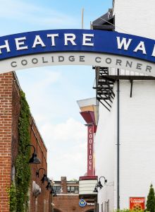 The image shows a storefront or entrance with a sign &ldquo;THEATRE WALK&rdquo; above a brick arch, a small sculpture or statue on the left, and a clear blue sky.