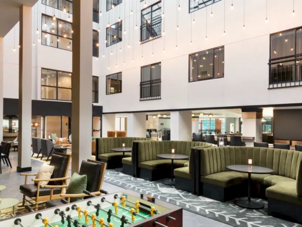 A modern hotel lobby with seating areas, a foosball table, and a bright open atrium with multiple windows and string lights, all in neutral tones.