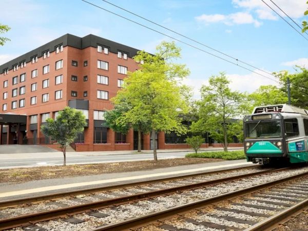 A modern brick hotel beside a tram line, with green trees, a sidewalk, and a tram approaching along railway tracks.